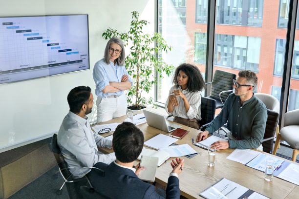 Senior female ceo and multicultural business people discussing company presentation at boardroom table. Diverse corporate team working together in modern meeting room office. Top view through glass